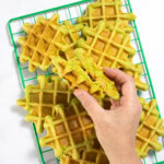 Green wire cooling rack with a bunch of spinach waffles stacked up with my hand holding one split into two halves over the waffles.