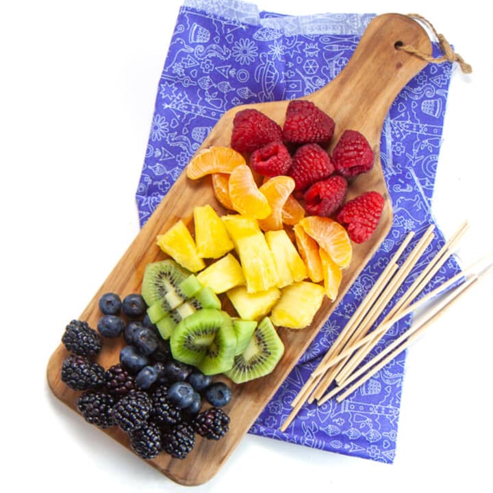 A small cutting board Full of rainbow colored fruit skewer sitting next to it in a purple kids napkin all against a white background.