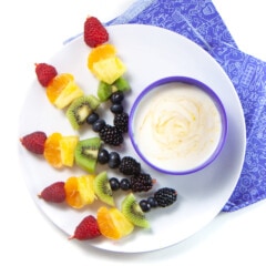 A white plate with several rainbow colored fruit skewers in a bowl of yogurt dip with a purple kids napkin against a white background.