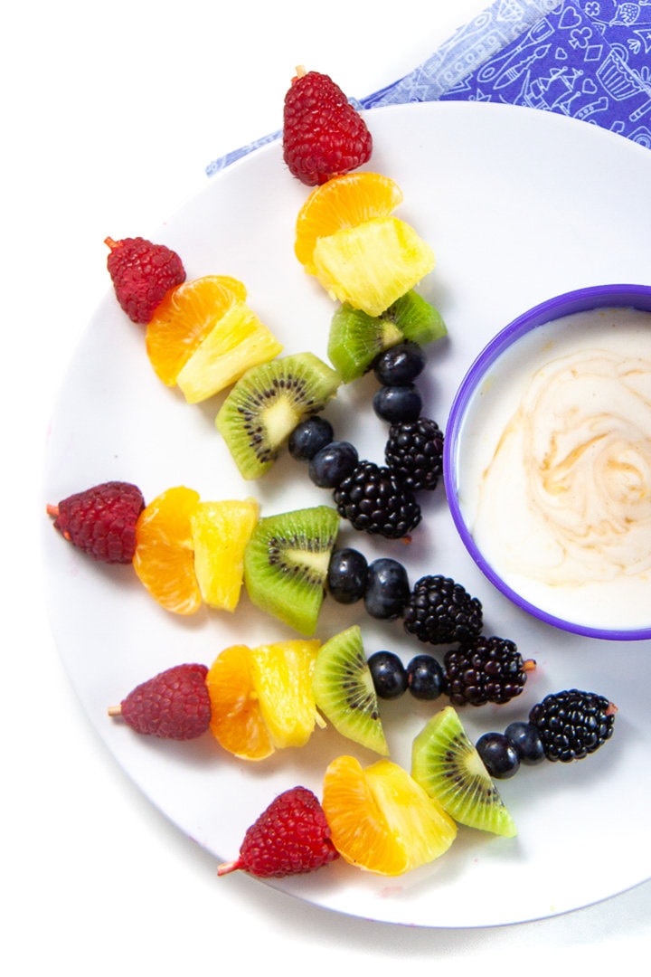 A white plate full of rainbow colored fruit skewers and a purple kids bowl full of lemon honey yogurt against a white background.