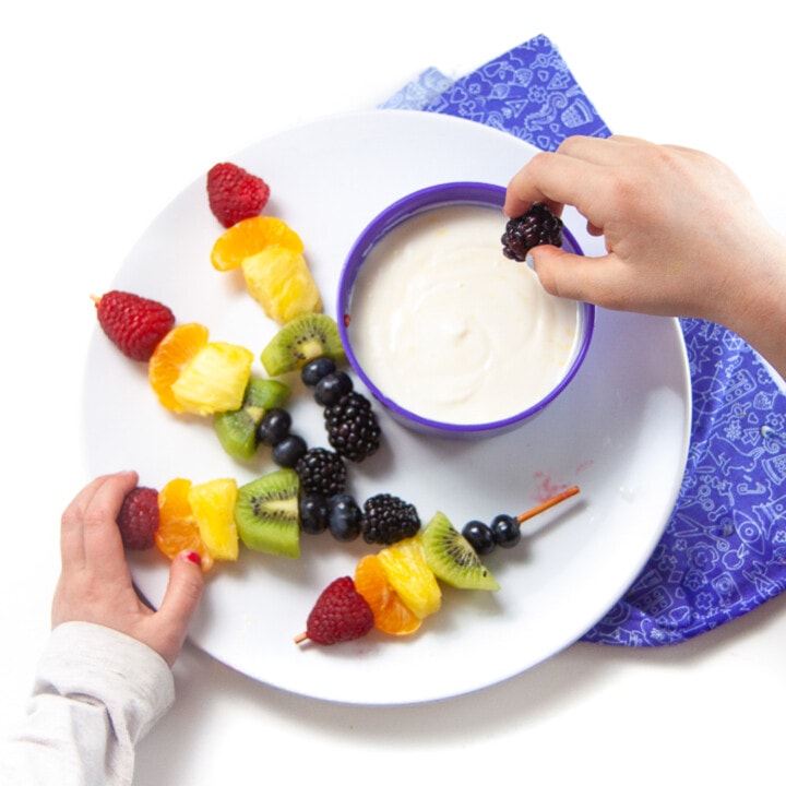 A white plate full of rainbow fruit skewers and a bowl of lemon honey yogurt dip with a small kids hand reaching for the skewer.