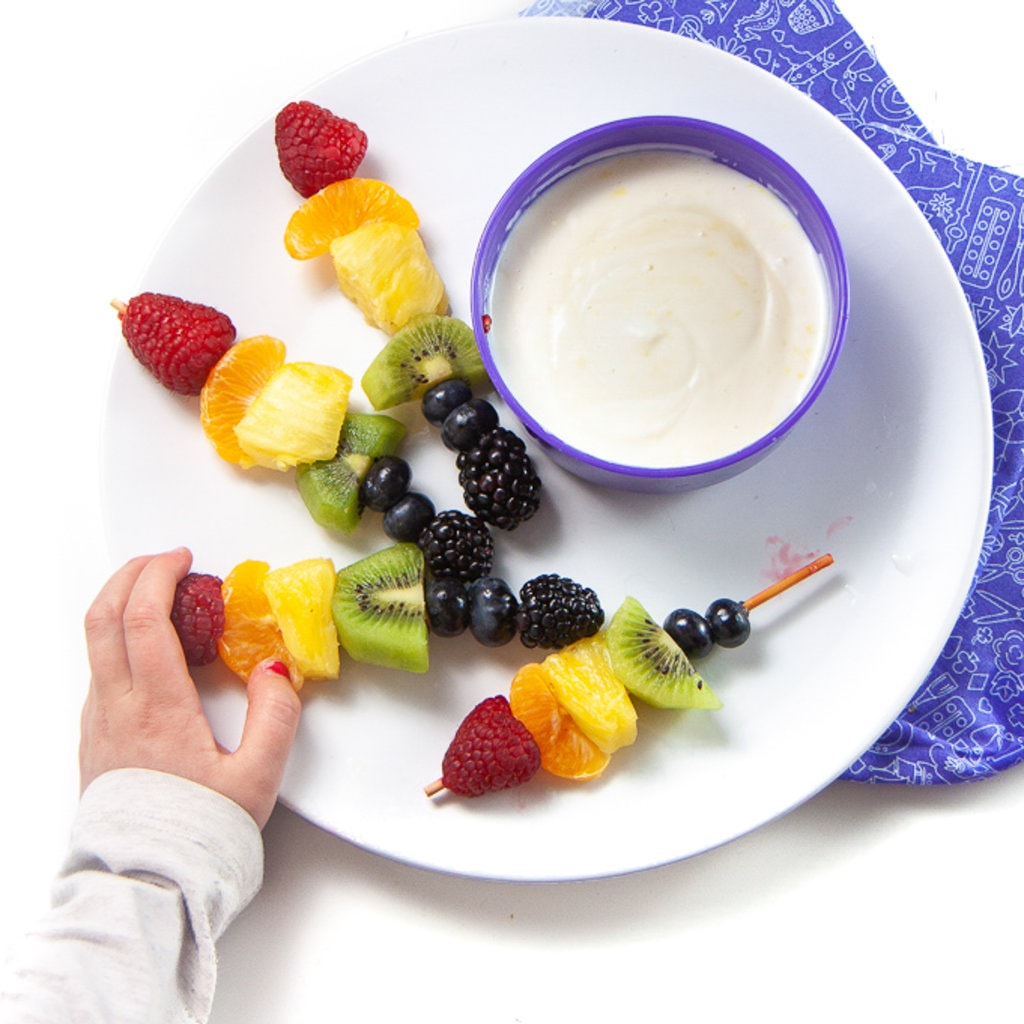A white plate full of rainbow fruit skewers and a bowl of lemon honey yogurt dip with a small kids hand reaching for the skewer.
