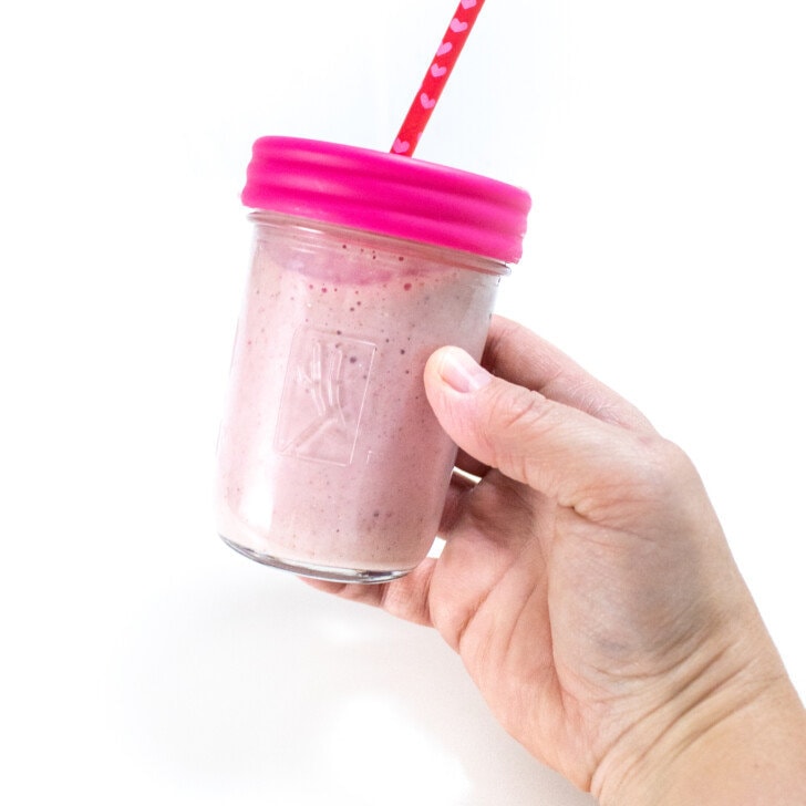 Hand holding a clear glass kids cup full of pink strawberry banana smoothie with a pink lid and pink straw.
