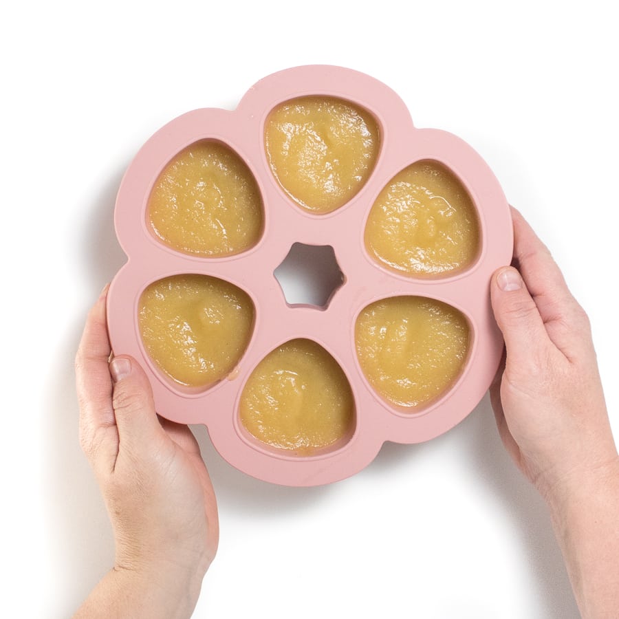 Pink baby food storage container with puréed apples and hands holding it.