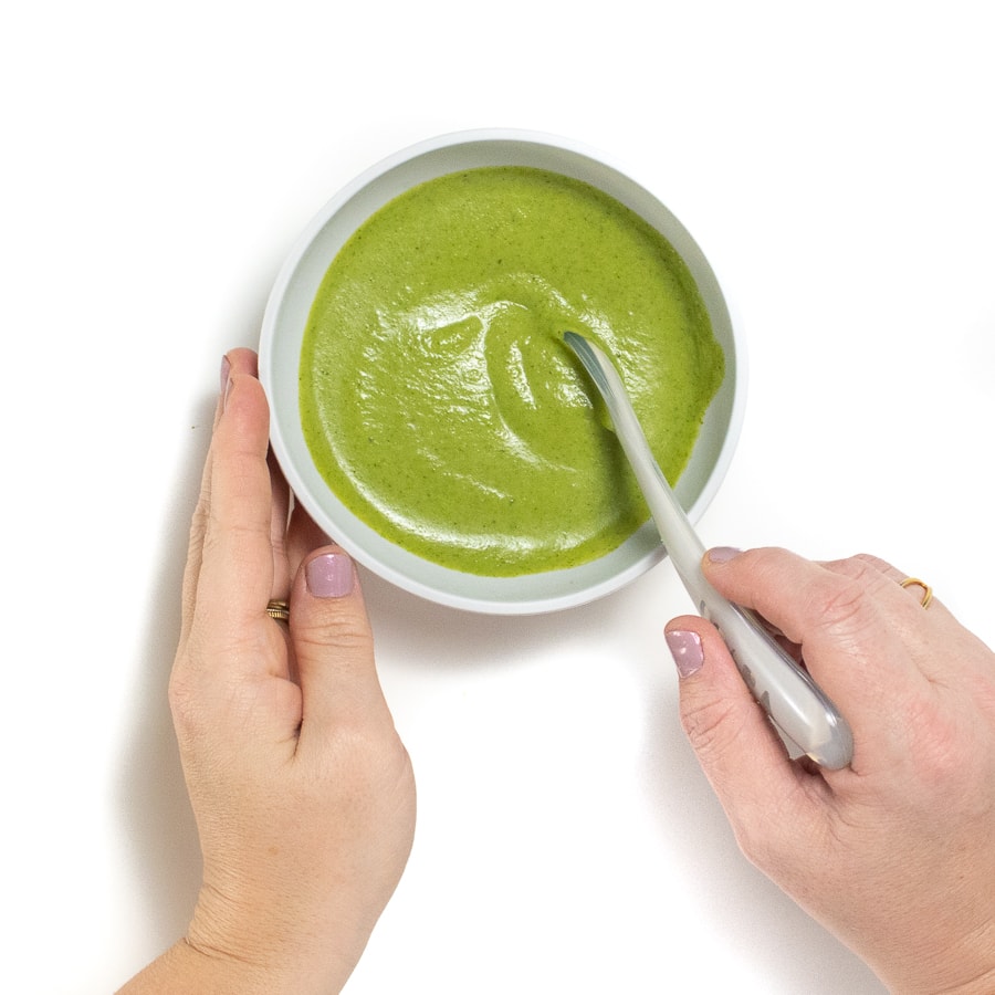 hands holding a grey baby bowl and stirring a smooth broccoli puree.