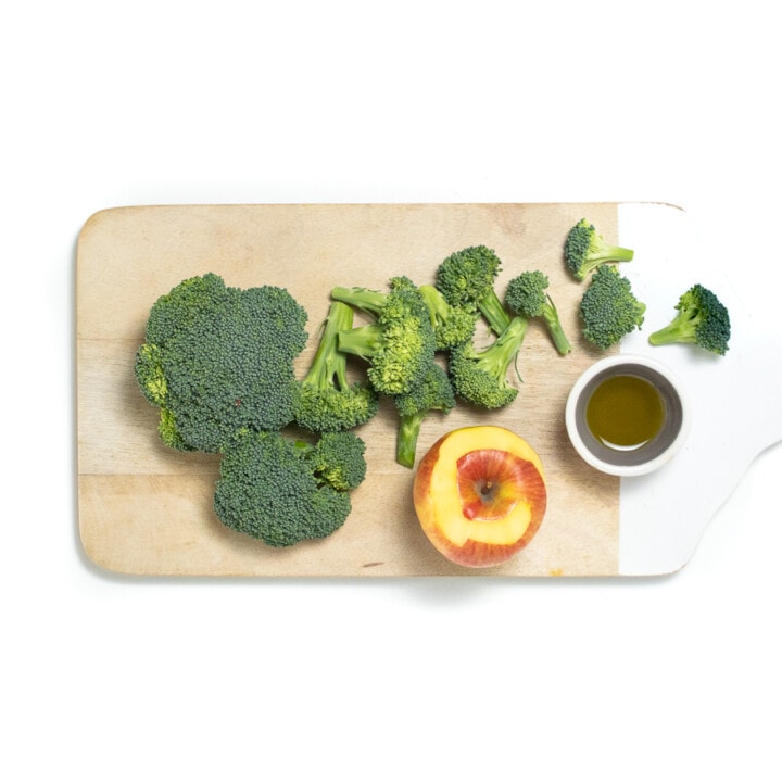 Cutting board with cut broccoli florets with a small bowl for olive oil and a half way peeled apple.