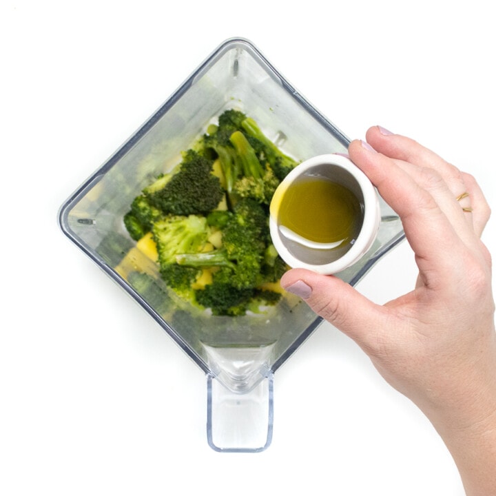 Blender with steamed broccoli with a hand pouring in olive oil.