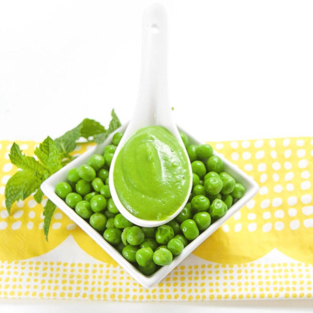 White spoon laying in a small dish full of peas on a yellow napkin with some mint on the napkin. In the spoon is a smooth pea puree. 