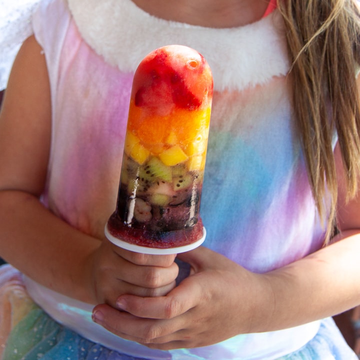 young girl holding a rainbow popsicle wearing a rainbow dress.