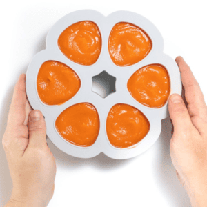 Hands holding a gray baby food storage tray filled with carrot puree over my white kitchen counter.
