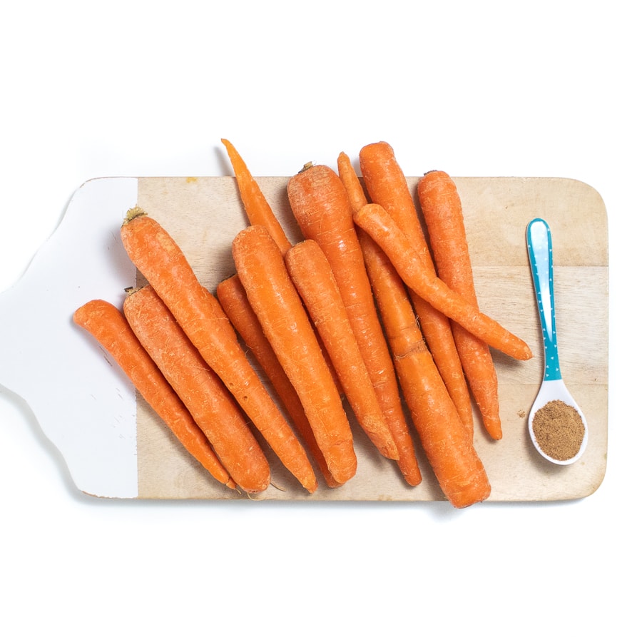 Wood cutting board with whole carrots in a pile and a blue spoon with nutmeg in it. 