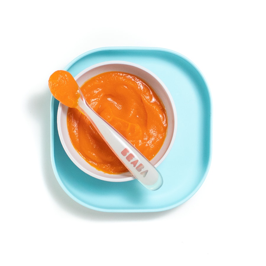 A blue play against a white background with a gray bowl with carrot purée and a gray spoon resting on top.