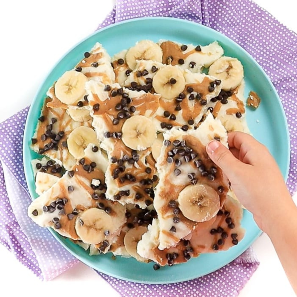 little kids hand holding a piece of banana yogurt bark over a kids plate full of bark that is resting on a purple napkin that is sitting on my white kitchen counter. 