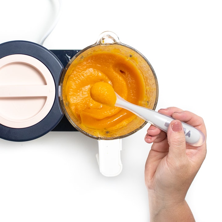 Hand holding a gray spoon over a jar full of baby food. 