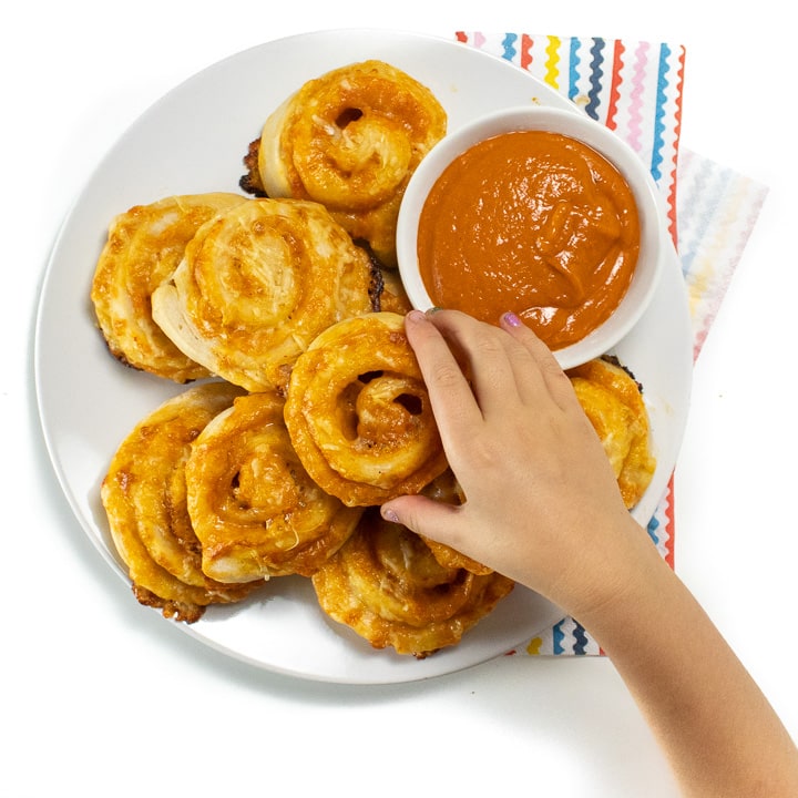 Hand reaching over a plate of veggie hidden pizza rollups with a side of sauce ready for kids to take and eat.