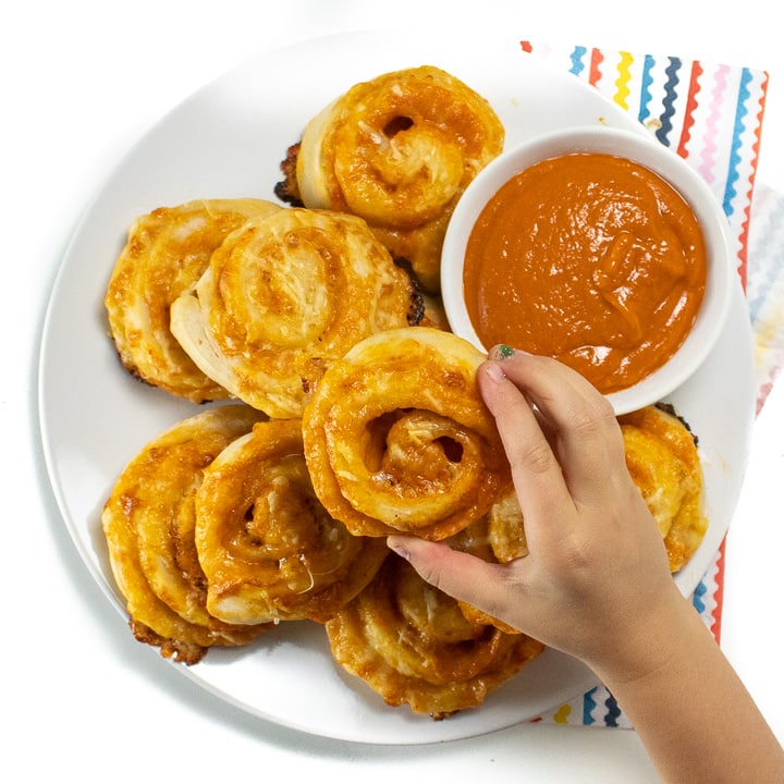 Hand reaching over a plate of veggie hidden pizza rollups with a side of sauce ready for kids to take and eat.