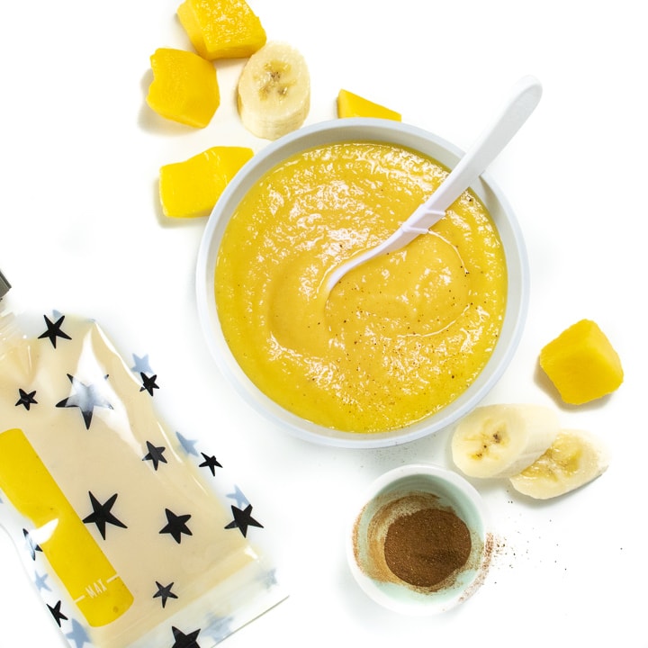 A small bowl of mango puree surrounded by fresh produce and a reusable baby food pouch. 