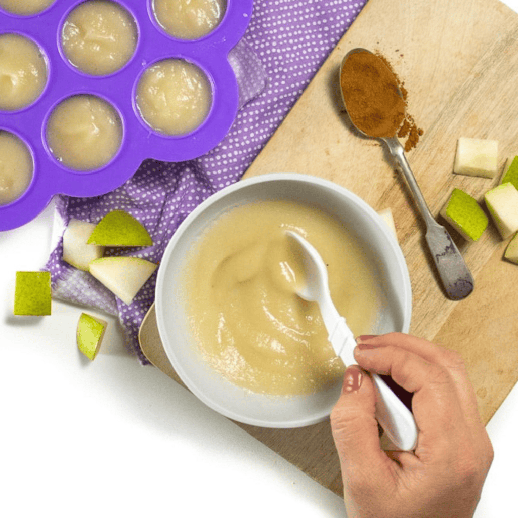 hand swirling a bowl full of smooth pear puree. The bowl is sitting on a cutting board with a spoonful of nutmeg and pieces of pear tossed around as well as a freezer container full of puree. All on my white kitchen counter.