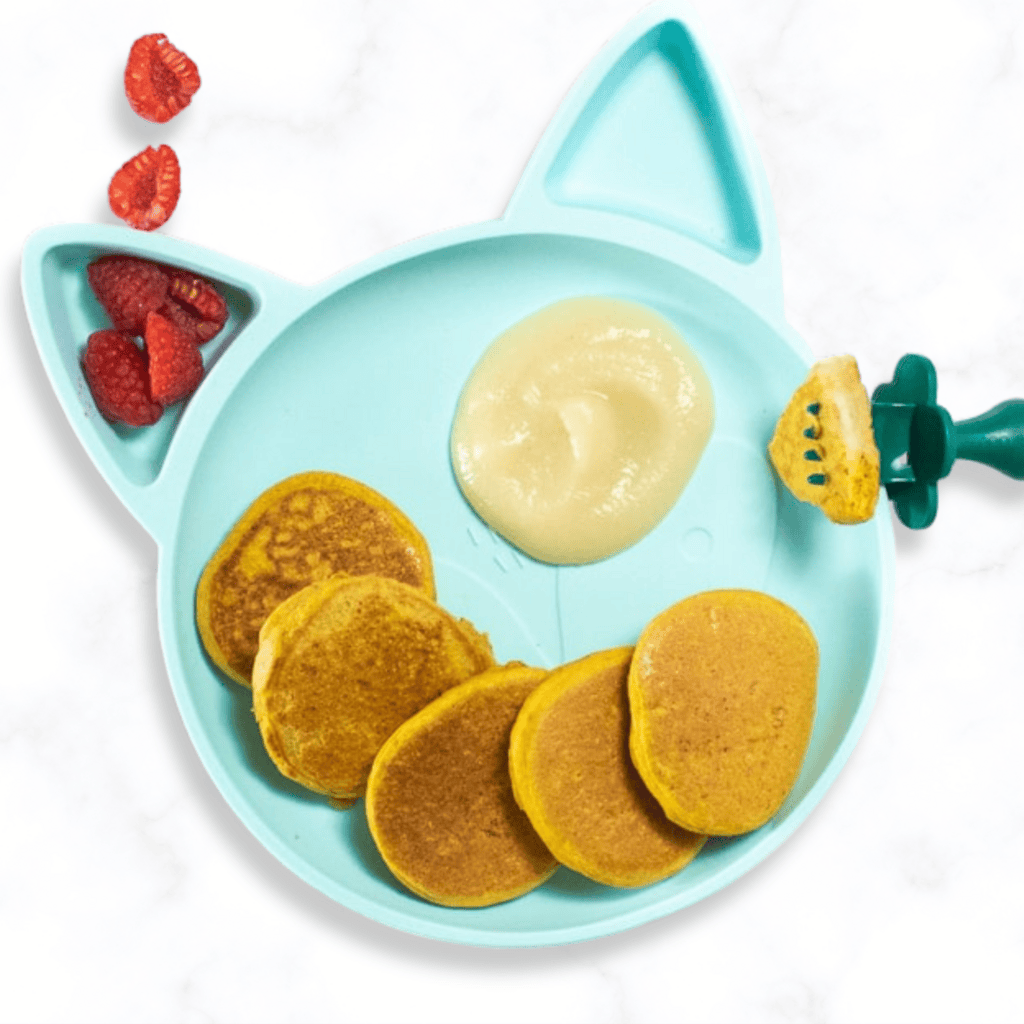 blue baby plate sitting on a kitchen counter with pumpkin pancakes, cut raspberries and a pear sauce for kids.