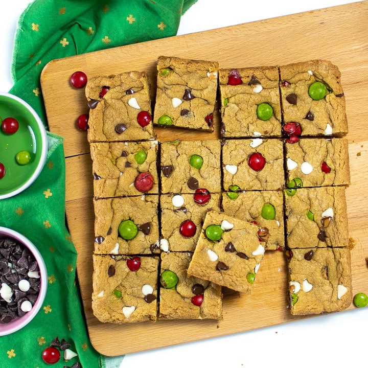 Christmas Cookie bars made with whole wheat flour sitting on a cutting board with dye-free candy and chocolate chips around it.