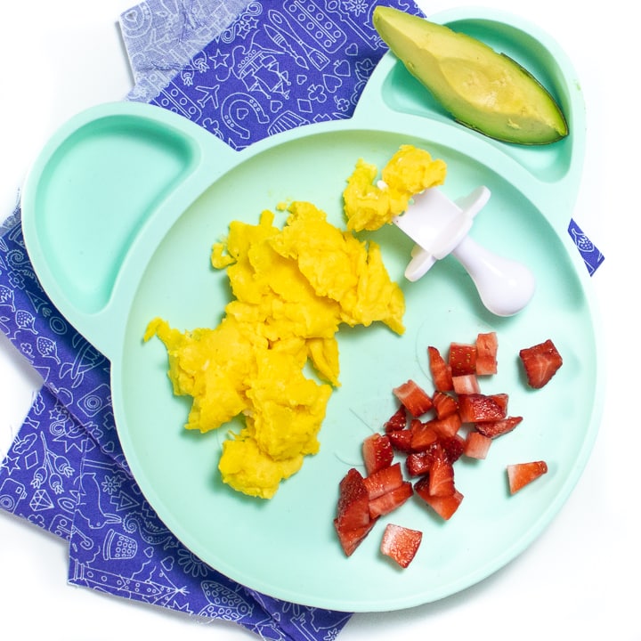 Teal plate on purple napkin with easy scrambled eggs, avocado and chopped strawberries.