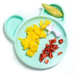 Teal plate with breakfast for baby - scrambled eggs, avocado and chopped strawberries.