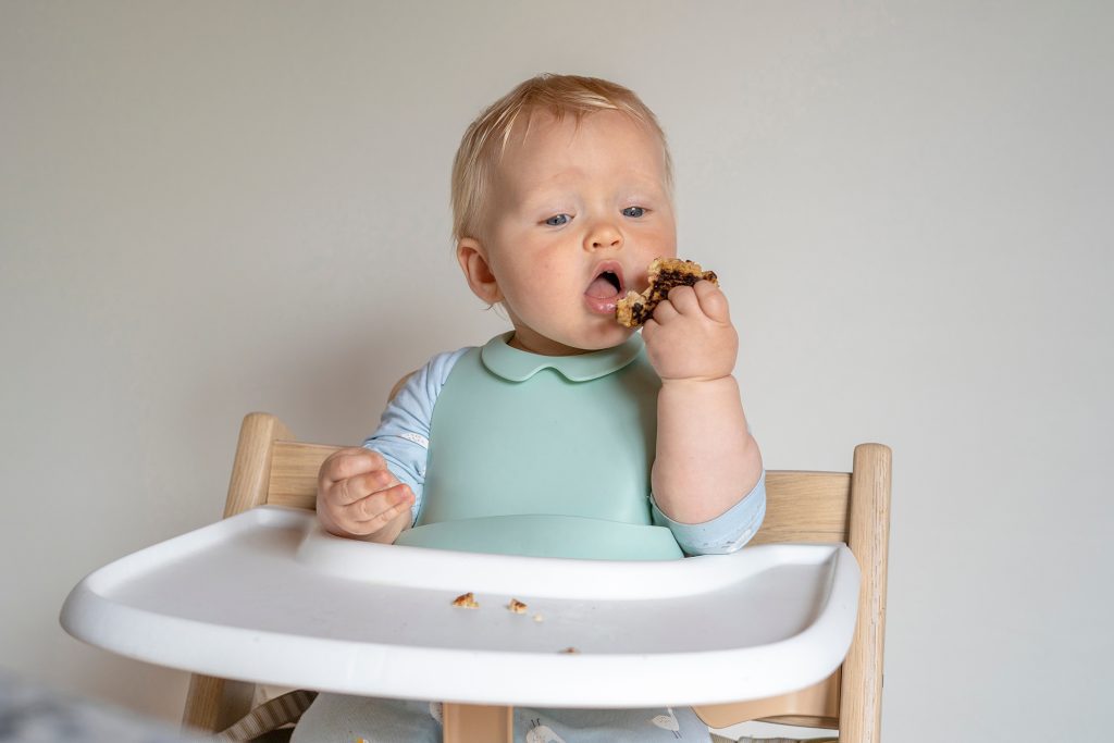 Baby sitting in a wood and white high chair feeding themselves a patty.
