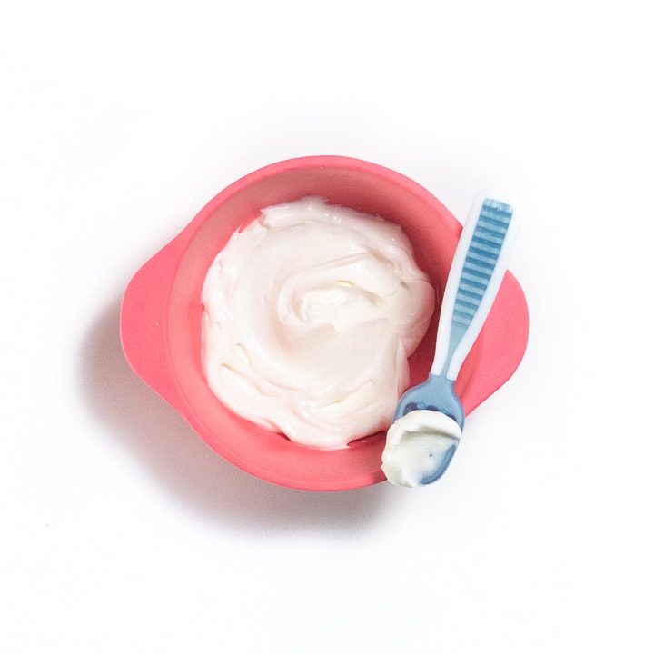 Baby spoon resting on a pink baby bowl with yogurt in it.