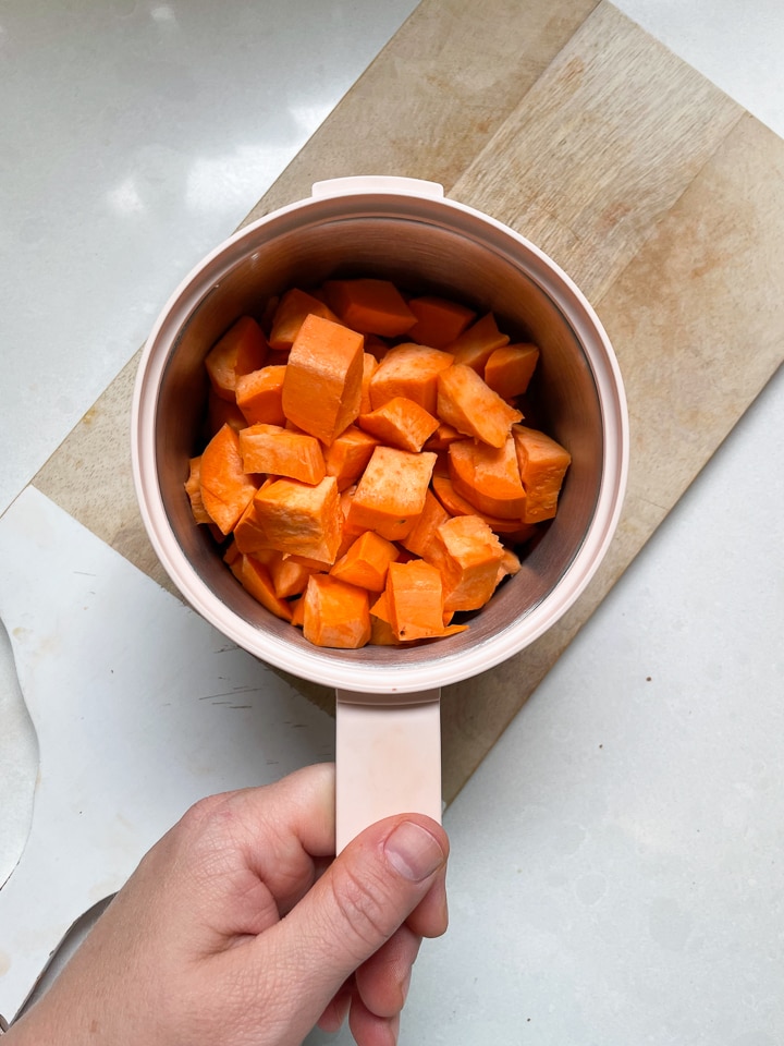 Hand holding the Beaba stainless steel basket with chopped sweet potatoes inside.