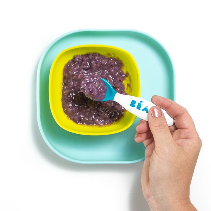Teal plate with green bowl with purple blueberry oatmeal inside with a hand holding a baby spoon with oatmeal on it.