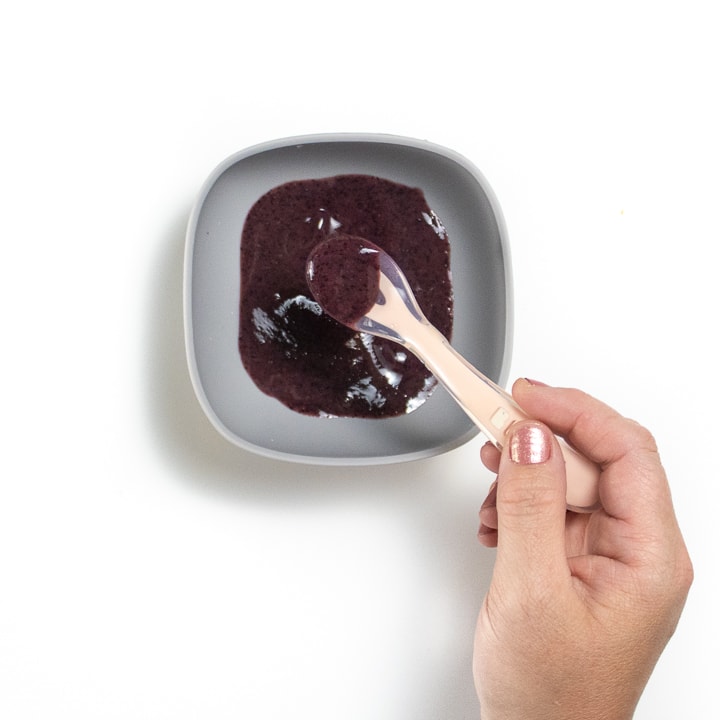 Hand holding a pink spoon with blueberry puree inside.