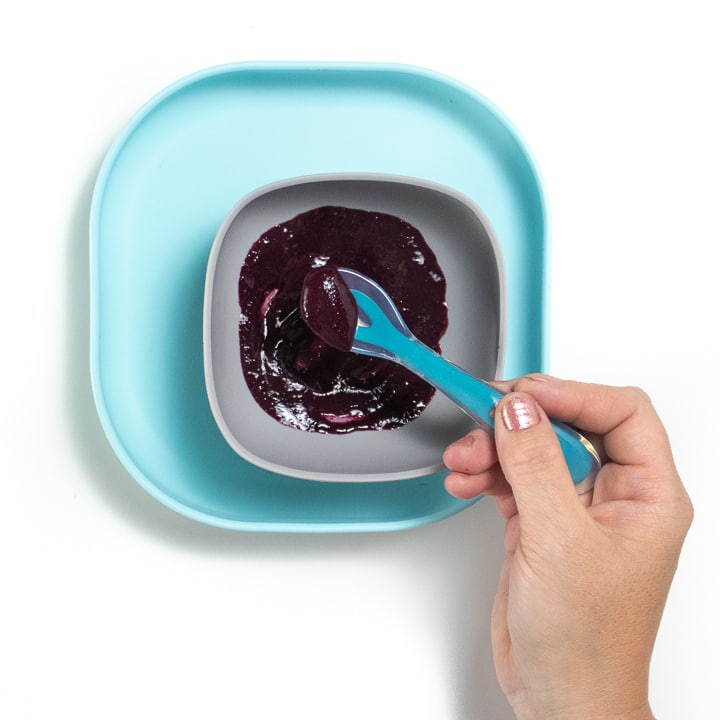 Teal baby plate with gray bowl with blueberry puree inside with hand holding spoon stirring puree.