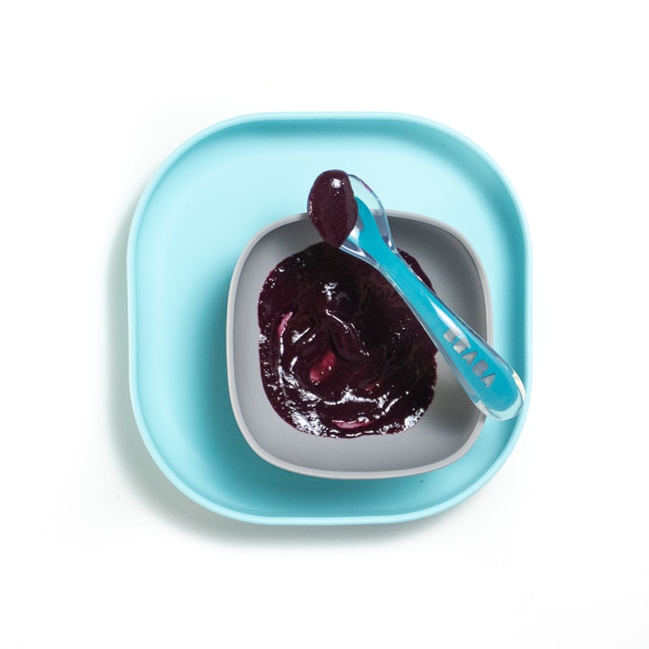 Teal baby plate with gray bowl with blueberry puree inside with darker blue spoon resting on bowl.