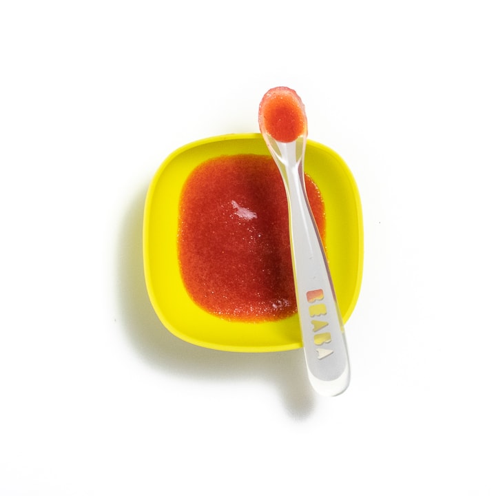 Yellow baby bowl with a pink strawberry puree inside with a spoon resting on the bowl.