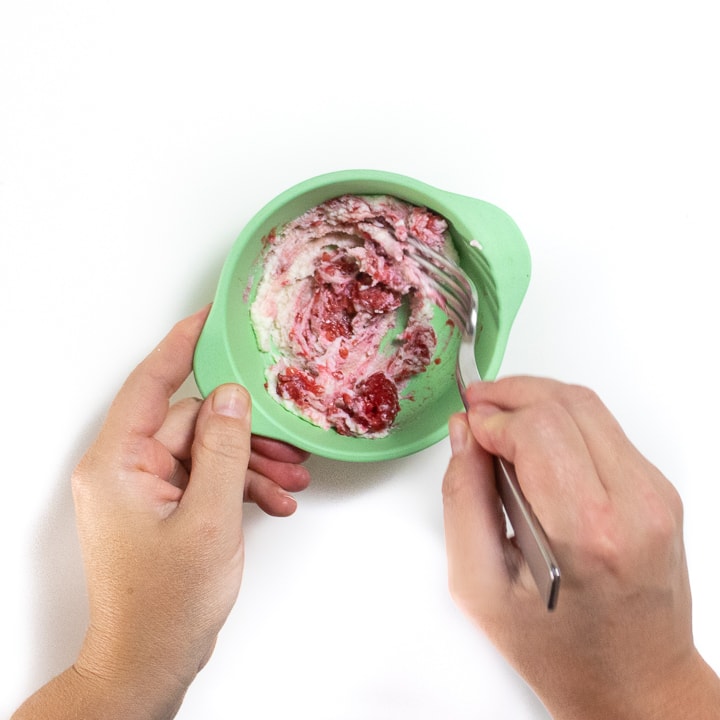 Hands holding and using a silver fork to mash raspberryies and ricotta in a green bowl.