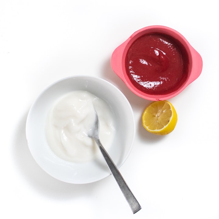 White bowl with yogurt, lemon and a pink bowl of raspberry puree on a white background.