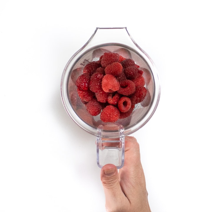 Hand holding a clear blender with fresh raspberries inside