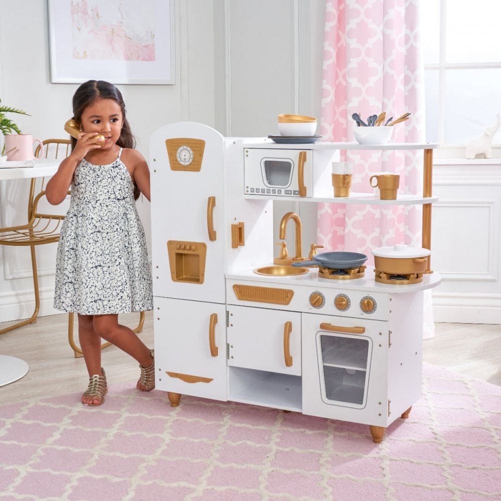 Young girl playing talking on a gold phone standing next to a white and gold play kitchen.