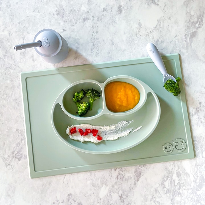 sage ezpz mat on a marbled counter with broccoli, peach puree and ricotta with raspberries, also have the mini cup and ezpz fork on the mat.