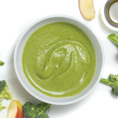 Gray baby bowl with a creamy and smooth broccoli puree in it with broccoli and apples scattered on a white background.