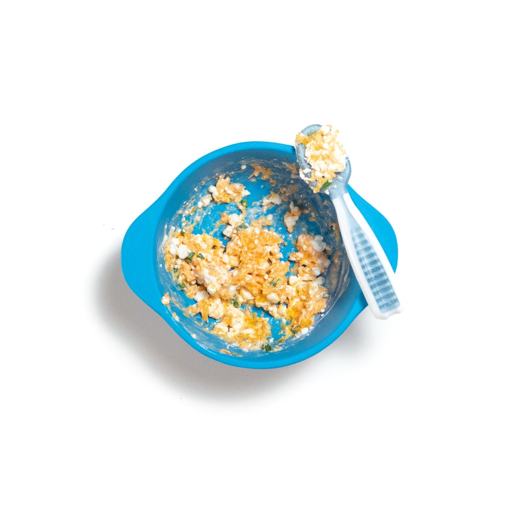 A blue baby bowl with mashed steamed carrots, cottage cheese and chives with a baby spoon rest in on the edge.