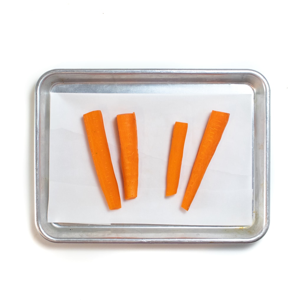 A silver baking tray with four sticks of carrots ready to be roasted.