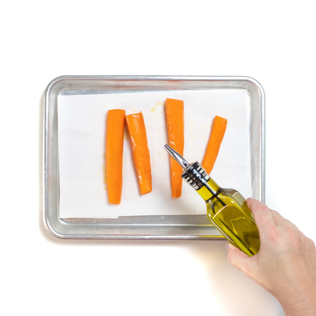 Is silver baking tray on a white background with four long strips of carrots with a hand pouring olive oil on top.