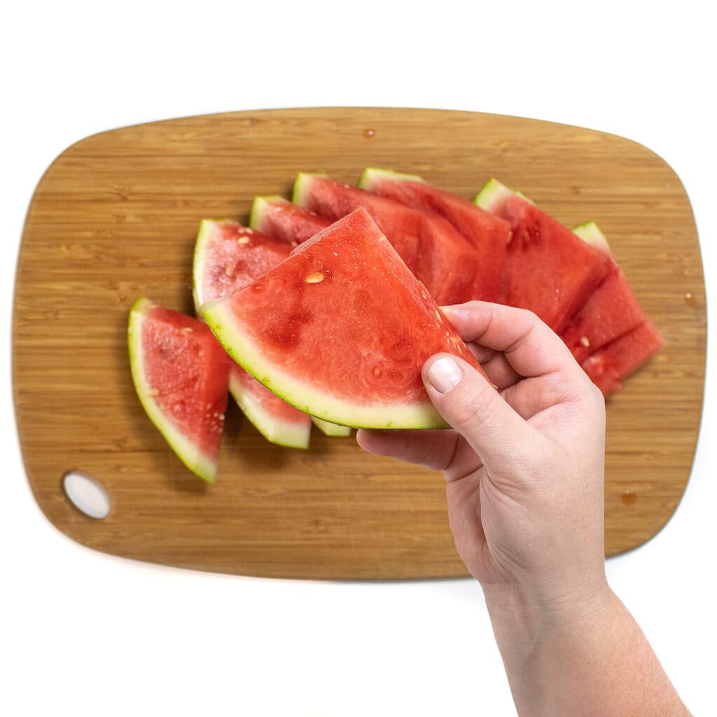 A hand holding a slice of watermelon over a wooden cutting board.