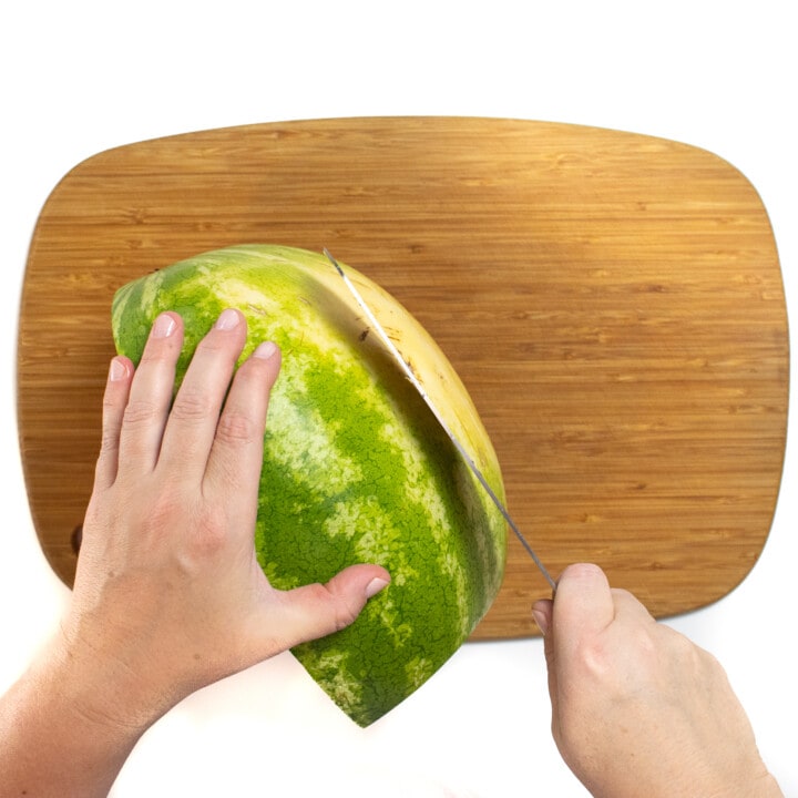 Wooden cutting board with a watermelon and a hand holding a knife cutting the bottom off of it.