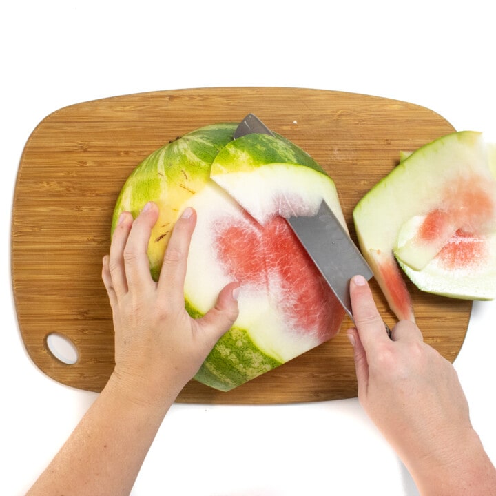 A wooden cutting board with a watermelon on its side with a hand trimming the rind off of the watermelon.