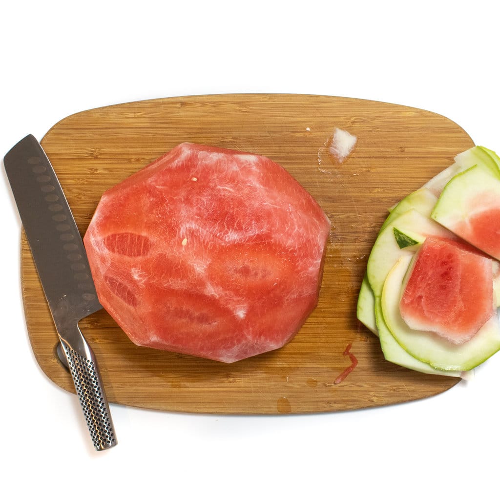 A board against a white background, with a knife, a half of a skin watermelon and the peels of a watermelon.
