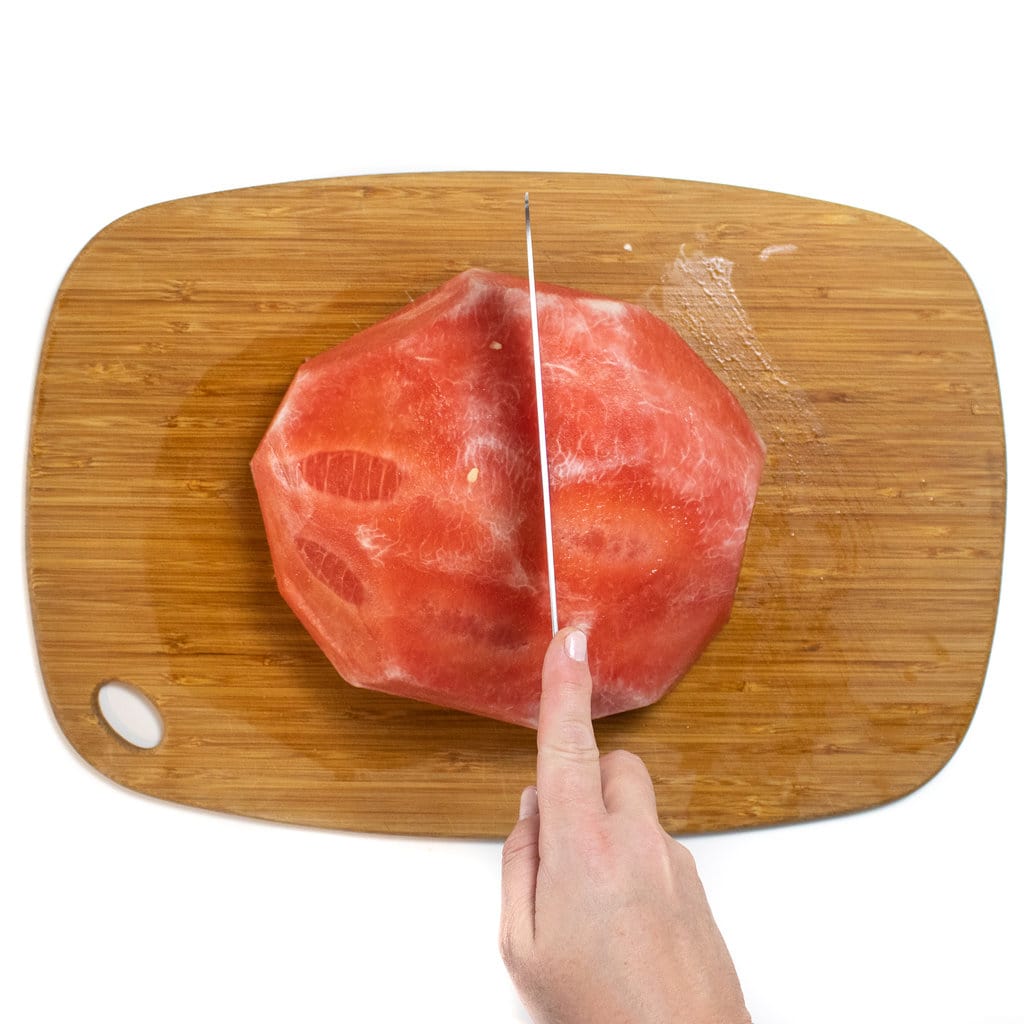 A half of a peeled watermelon on a wooden cutting board with a hand holding a knife cutting it in half.