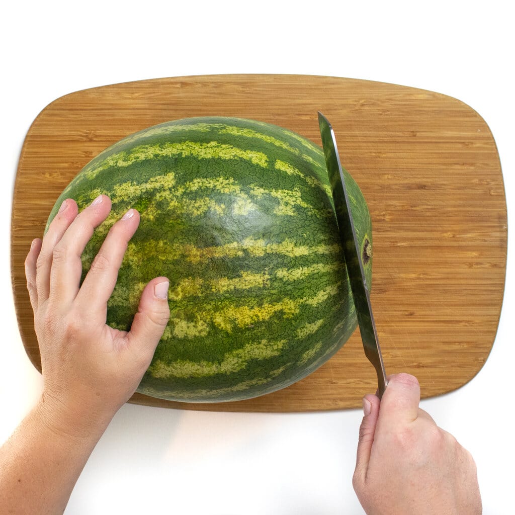 A watermelon on a cutting board with hands cutting off one end.