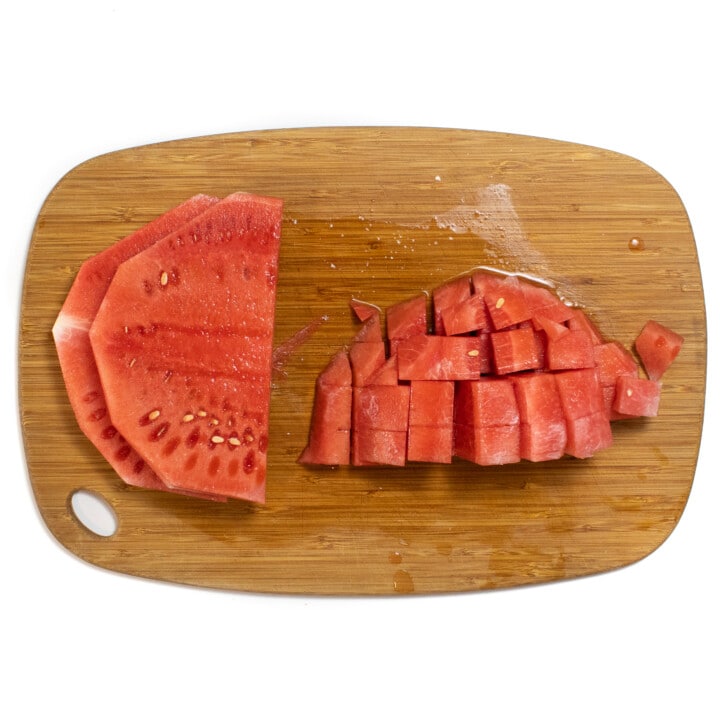 A wooden cutting board with two thick slices of watermelon in a pile of cubes of watermelon